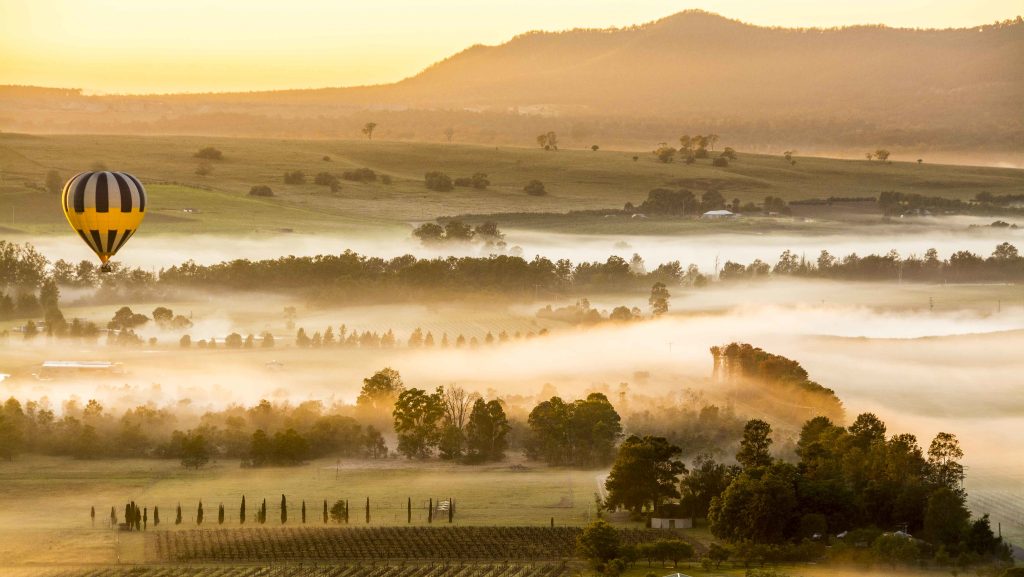 Early morning hot air balloon over the Hunter Valley with morning mist and yellow lighting the vineyard fields.