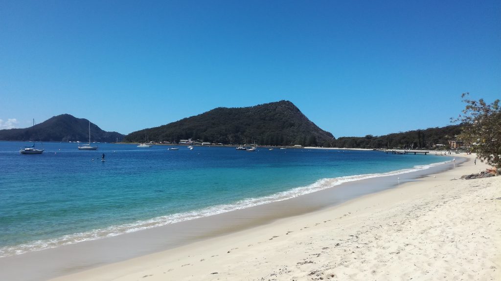 A scenic view of Shoal Bay, NSW, Australia, featuring pristine white sands, a clear blue sky, and lush mountains in the background, ideal for hiking.