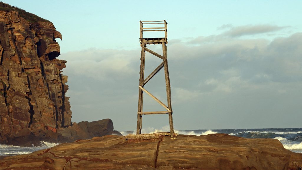 The Redhead Shark Tower and its iconic rock glow warmly in the afternoon sun. The tower, a wooden relic of the past, stands slightly shorter than the towering rock cliff, adding a touch of history to the scenic coastal landscape.