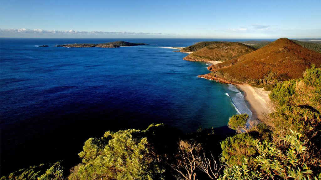 A scenic view of the Tomaree Peninsula from the headland, showcasing the beaches and rugged coastline below, with an island visible in the distance, separated from the mainland by a narrow channel.