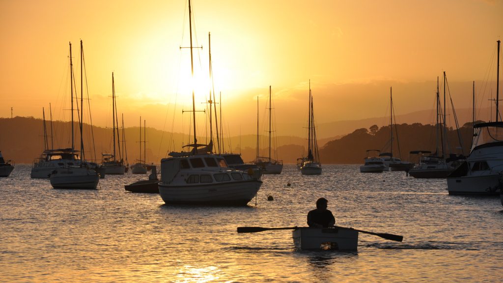 Rower gliding through sailing boats against a sunset-lit backdrop.